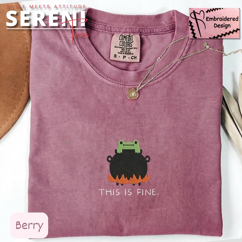 May include: Berry-colored t-shirt with a crew neck. The shirt features an embroidered design of a frog on a cauldron with the text "THIS IS FINE." A silver necklace with a pendant is also visible.