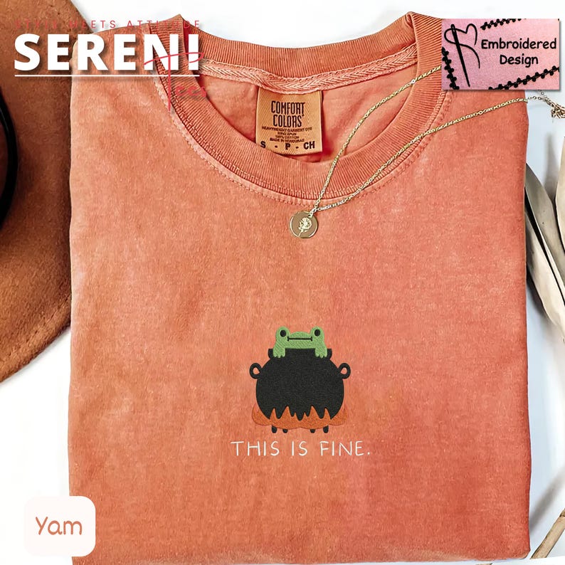 May include: A coral-colored t-shirt with a round neckline. The shirt features an embroidered design of a frog on a cauldron with the text "THIS IS FINE." A gold necklace with a pendant is draped on the shirt.