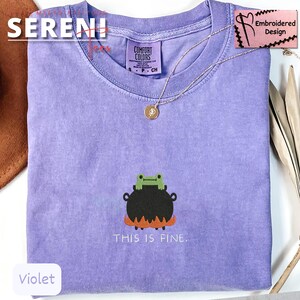 May include: Violet-colored t-shirt with a frog-in-cauldron graphic and the text "THIS IS FINE." The shirt has a crew neck and short sleeves. A gold-colored necklace with a pendant is draped on the shirt. The shirt is labeled "Comfort Colors."