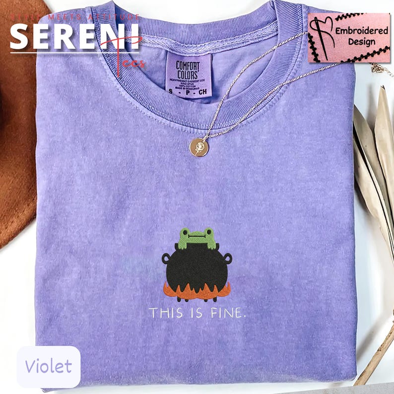 May include: Violet-colored t-shirt with a frog-in-cauldron graphic and the text "THIS IS FINE." The shirt has a crew neck and short sleeves. A gold-colored necklace with a pendant is draped on the shirt. The shirt is labeled "Comfort Colors."
