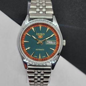 Pode incluir: Um relógio de pulso automático Seiko prateado com mostrador verde-azulado e anel externo vermelho. O relógio apresenta ponteiros e marcadores dourados e um visor de dia e data. A pulseira do relógio é feita de elos de metal prateado.