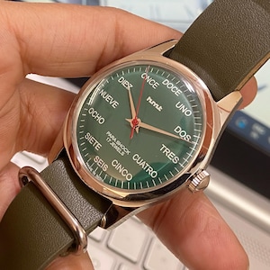 Puede incluir: Un reloj de pulsera con esfera verde y caja dorada. La esfera muestra la hora con agujas doradas y n&uacute;meros espa&ntilde;oles. El reloj tiene una correa de cuero verde y el texto "PARA SHOCK 17 JEWELS".