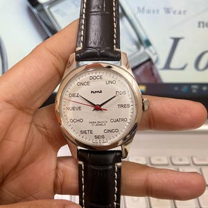 May include: A silver wristwatch with a black leather strap. The watch face has a white background with black hands and black numbers in Spanish. The watch face also has the words "humt" and "Para Shock 17 Jewels".