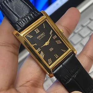 May include: A rectangular Seiko Quartz watch with a gold-tone case and a black dial. The dial features gold Roman numerals, hands, and the Seiko logo. The watch has a black leather band.