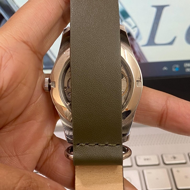 Puede incluir: Un reloj de pulsera plateado con una correa de cuero verde. La esfera del reloj es visible, mostrando los intrincados mecanismos internos. La correa est&aacute; sujeta con herrajes plateados.