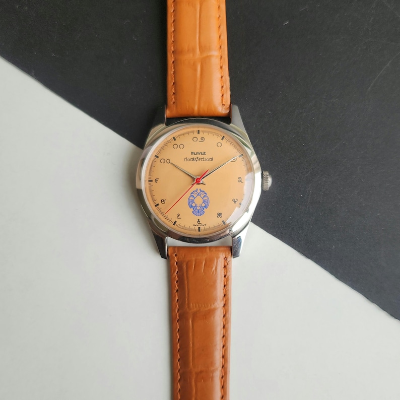 K&ouml;nnte beinhalten: Eine Armbanduhr mit hellbraunem Lederarmband und silberfarbenem Geh&auml;use. Das Zifferblatt ist hellpfirsichfarben mit schwarzen Ziffern und dem Text "hmt Hook&rood". Die Uhr hat einen roten Sekundenzeiger und ein blaues Emblem.