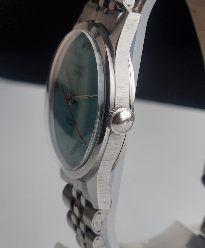 May include: A silver wristwatch with a green face and red hands. The watch has a clear, domed crystal and a silver band. The watch face has the word "TIME" printed on it. The watch is a classic style.