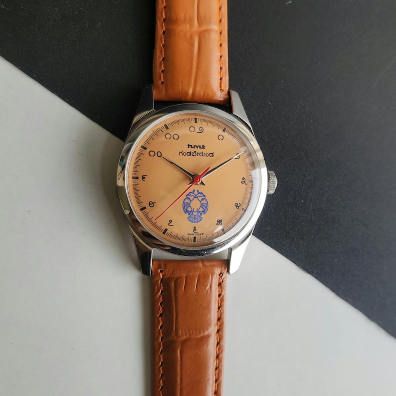 K&ouml;nnte beinhalten: Eine Vintage-Armbanduhr mit hellbraunem Zifferblatt, silbernem Geh&auml;use und einem braunen Lederarmband. Das Zifferblatt hat schwarze Ziffern, rote und schwarze Zeiger und ein blaues Emblem. Das Armband ist aus strukturiertem Leder im Krokodil-Stil.