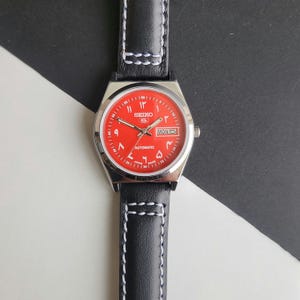 May include: A stainless steel Seiko 5 automatic watch with a vibrant red dial and black leather strap. The watch face features Arabic numerals and a day-date display. The black strap has white stitching.