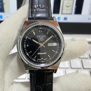 Puede incluir: Un reloj de pulsera automático Seiko 5 de esfera negra con caja plateada y correa de cuero negra. La esfera del reloj tiene marcadores de hora y agujas plateadas, una ventana de fecha que muestra "MON 17", y el texto "SEIKO 5 Automatic" y "JAPAN MADE".