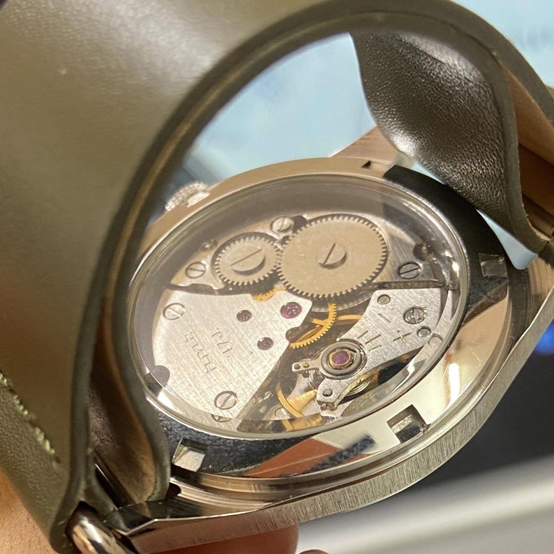 Puede incluir: Primer plano de un reloj de pulsera con la parte trasera transparente, que muestra el intrincado movimiento mec&aacute;nico. El reloj presenta una caja de metal plateado y una correa de cuero verde. Los engranajes internos, las joyas y los muelles son visibles.