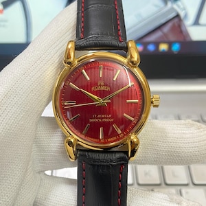Peut inclure: Une montre-bracelet dorée avec un cadran rouge et un bracelet en cuir noir. Le cadran de la montre a des aiguilles et des marqueurs dorés, et le texte "RW ROAMER 17 JEWELS SHOCK PROOF" est imprimé en blanc. Le bracelet a un détail de couture rouge.