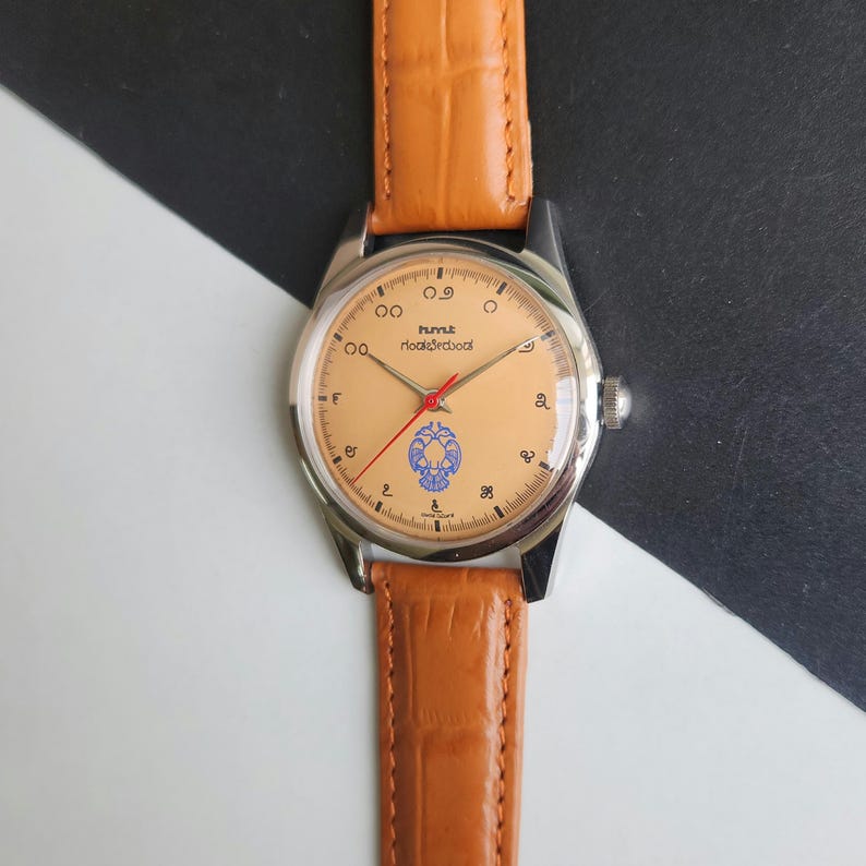 K&ouml;nnte beinhalten: Eine Vintage-Armbanduhr mit hellorangefarbenem Zifferblatt, silbernem Geh&auml;use und braunem Lederarmband. Das Zifferblatt der Uhr hat ein blaues Emblem und rote Zeiger. Das Armband hat eine hellbraune Farbe.