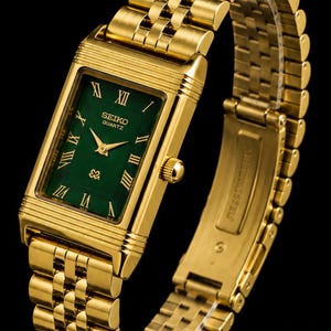 May include: A gold-tone rectangular Seiko Quartz watch with a deep green dial featuring gold Roman numeral hour markers and gold hands. The timepiece includes a matching gold-tone metal link bracelet with a secure clasp mechanism.