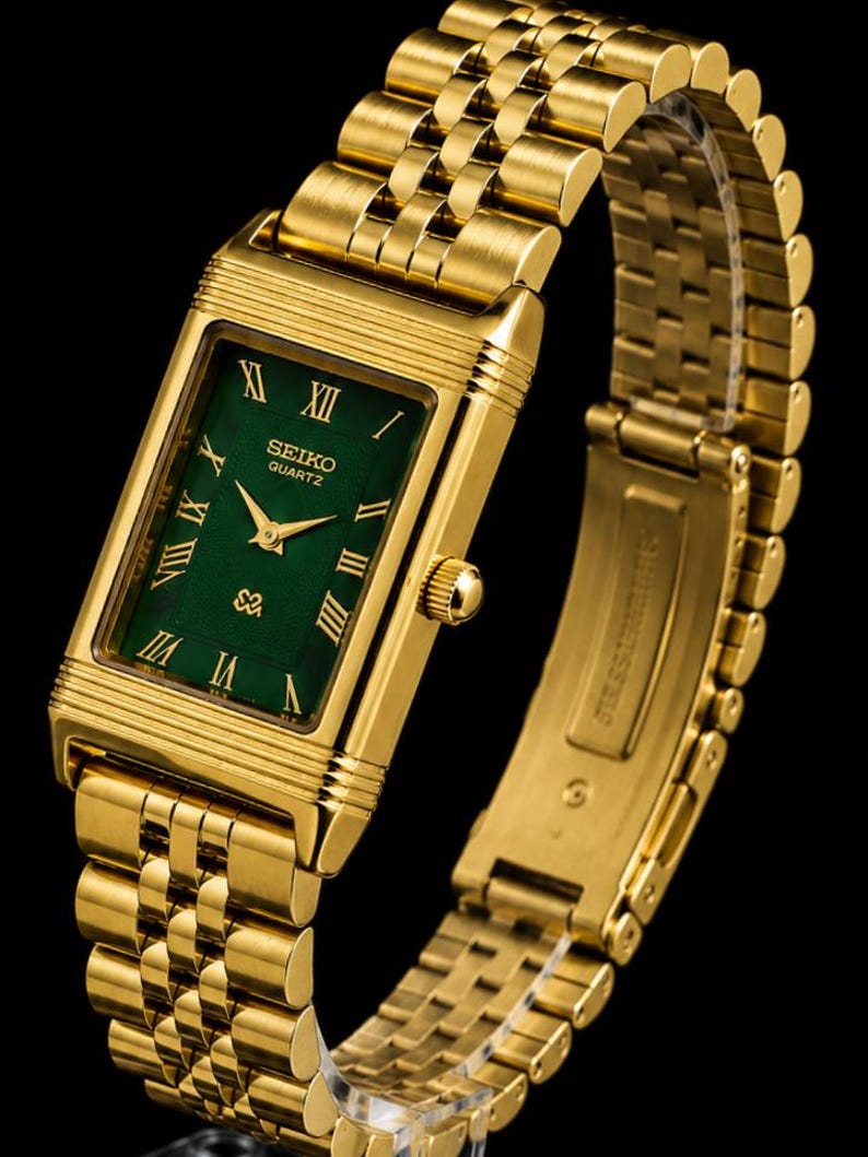 May include: A gold-tone rectangular Seiko Quartz watch with a deep green dial featuring gold Roman numeral hour markers and gold hands. The timepiece includes a matching gold-tone metal link bracelet with a secure clasp mechanism.