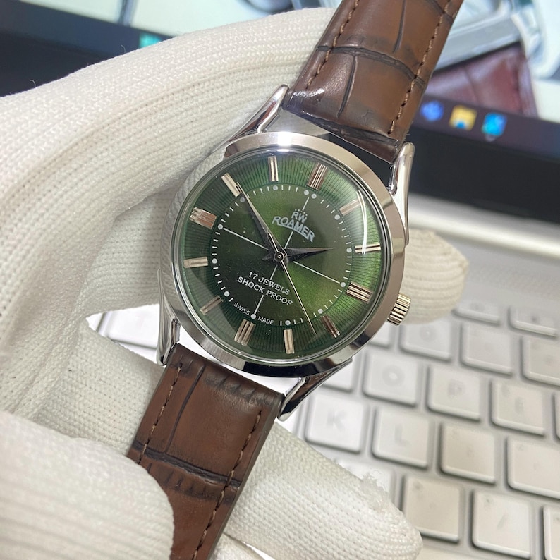 May include: A silver men's wristwatch with a green face and brown leather band. The watch face has gold accents and the text "17-JEWELS SHOCK PROOF" and "ROAMER" printed on it.