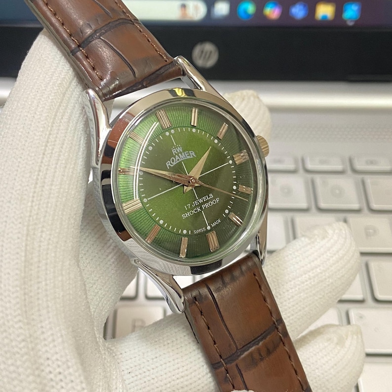 May include: A vintage Roamer wristwatch with a green face and silver casing. The watch has a brown leather band and says "RW Roamer" and "17 Jewels Shock Proof" on the face.