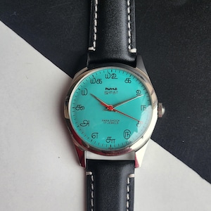 May include: A vintage wristwatch with a teal face and silver casing. The watch has black leather straps with white stitching. The watch face displays text in an unfamiliar script, with red hands and a second hand.