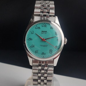 May include: Silver-toned wristwatch with a turquoise face and a silver band. The watch face features black markings, red hands, and text in a foreign language. The watch is labeled "PARA SHOCK 17 JEWELS" and "INDIA".