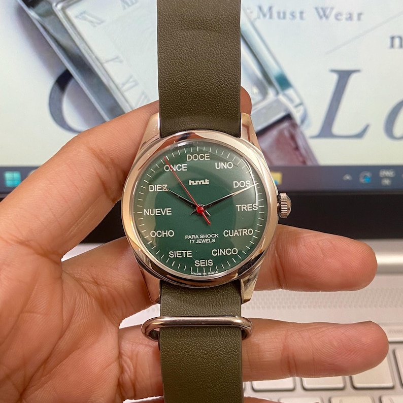 Puede incluir: Un reloj de pulsera con esfera verde y correa de cuero verde. La esfera muestra n&uacute;meros en ingl&eacute;s y espa&ntilde;ol. El reloj tiene una caja plateada y un segundero rojo. El texto "hmt" es visible en la esfera.
