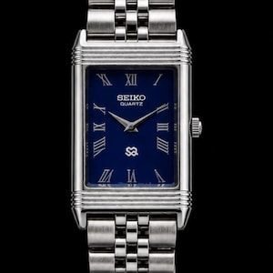 Może przedstawiać: Prostokątny zegarek Seiko Quartz z głęboką niebieską tarczą, srebrnymi rzymskimi cyframi i srebrnymi wskazówkami. Zegarek posiada metalową bransoletę z ogniw i srebrną kopertę z poziomymi żłobieniami na górze i na dole tarczy.
