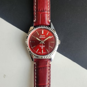 May include: A vintage wristwatch with a deep red face and a silver bezel. The watch has a matching red leather strap with white stitching. The watch face displays the brand name and the words "PARA SHOCK 17 JEWELS".