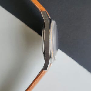 K&ouml;nnte beinhalten: Seitenansicht einer silbernen Armbanduhr mit einem orangefarbenen Lederarmband. Das Zifferblatt ist sichtbar, mit einem klaren Kristall und einem hellorangefarbenen Innenraum. Das Armband ist etwa 2 cm breit.