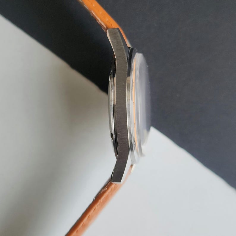 K&ouml;nnte beinhalten: Seitenansicht einer silbernen Armbanduhr mit einem orangefarbenen Lederarmband. Das Zifferblatt ist sichtbar, mit einem klaren Kristall und einem hellorangefarbenen Innenraum. Das Armband ist etwa 2 cm breit.