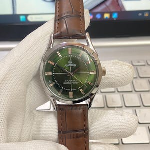 May include: A silver and green wristwatch with a brown leather band. The watch face has Roman numeral hour markers and the text "RW ROAMER 17 JEWELS SHOCK PROOF SWISS MADE".
