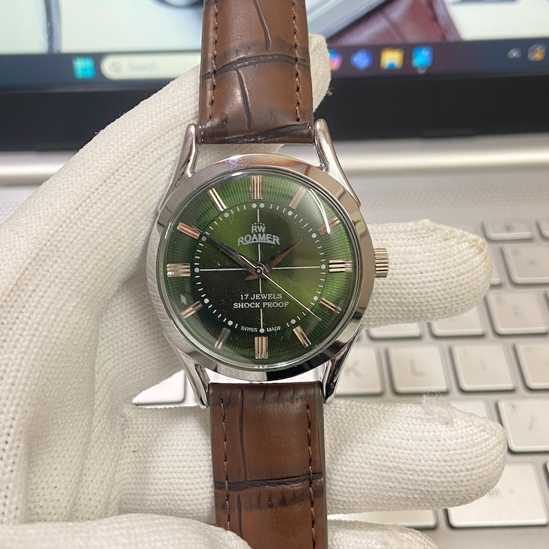May include: A silver and green wristwatch with a brown leather band. The watch face has Roman numeral hour markers and the text "RW ROAMER 17 JEWELS SHOCK PROOF SWISS MADE".