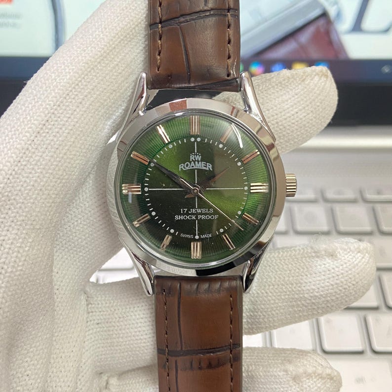 May include: A silver wristwatch with a green face and brown leather band. The watch face has Roman numeral hour markers and the text "RW ROAMER 17 JEWELS SHOCK PROOF SWISS MADE".