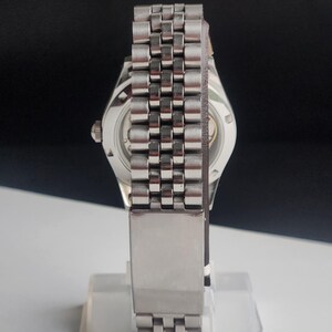 May include: A silver-toned wristwatch with a stainless steel band. The watch has a round face with a visible mechanism on the back. The band is made of small, connected rectangular links. The watch is displayed on a clear stand.
