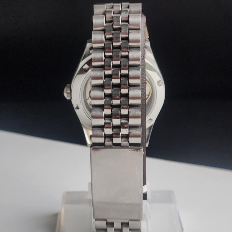 May include: A silver-toned wristwatch with a stainless steel band. The watch has a round face with a visible mechanism on the back. The band is made of small, connected rectangular links. The watch is displayed on a clear stand.