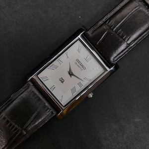 Puede incluir: Un reloj de pulsera rectangular Seiko Quartz con esfera blanca, números romanos negros, manecillas negras y un logotipo. El reloj está sujeto a una correa de cuero marrón oscuro con un patrón texturizado de imitación de cocodrilo.