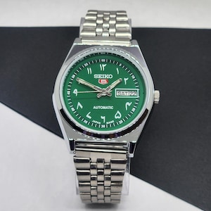 Puede incluir: Un reloj de pulsera automático Seiko plateado con esfera verde. La esfera muestra números arábigos, el día y la fecha. El reloj tiene una correa plateada y está sobre un fondo blanco y negro.