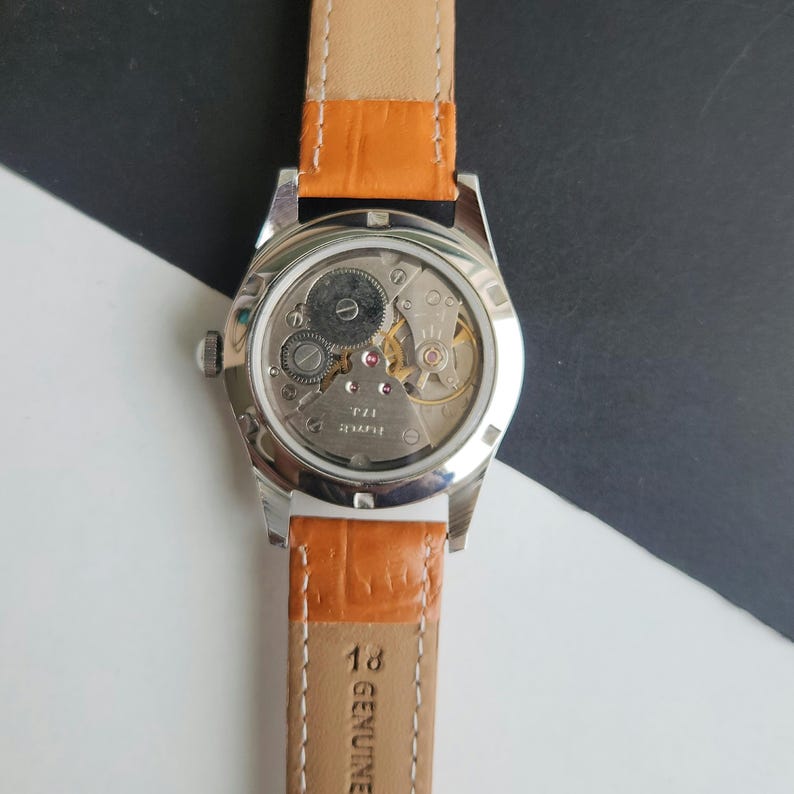 K&ouml;nnte beinhalten: Eine Vintage-Armbanduhr mit silbernem Geh&auml;use und einem orangefarbenen Lederarmband. Die R&uuml;ckseite der Uhr ist offen und zeigt das komplizierte mechanische Uhrwerk. Das Armband ist mit "GENUINE" und "18" gekennzeichnet.