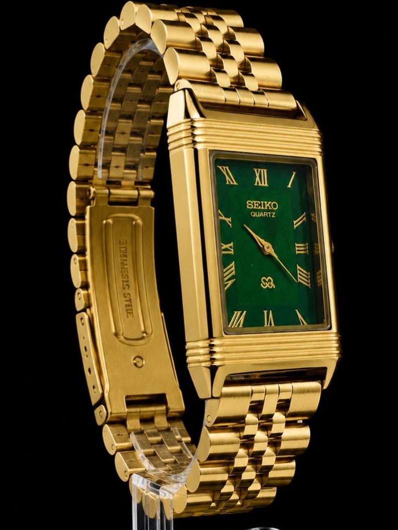 May include: A gold-tone rectangular wristwatch featuring a vibrant green dial with gold Roman numeral hour markers. The watch includes a matching gold-tone metal link bracelet and displays the text SEIKO QUARTZ on the face. The clasp is engraved with STAINLESS STEEL.