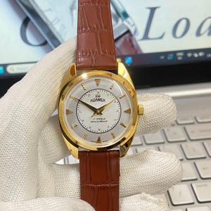 Peut inclure: Une montre-bracelet dorée avec un cadran blanc et un bracelet en cuir marron. Le cadran de la montre a des index horaires romains noirs et une aiguille des secondes noire. Le cadran de la montre porte également les inscriptions "ROAMER" et "17 JEWELS SHOCK PROOF".