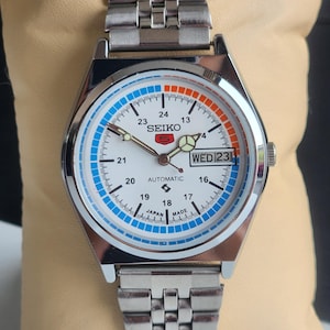 May include: A silver Seiko automatic wristwatch with a white face, blue and orange accents, and a day and date display. The watch face reads "WED 23".