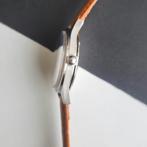 K&ouml;nnte beinhalten: Eine silberne Armbanduhr mit einem braunen Lederarmband. Das Zifferblatt der Uhr ist rund und das Armband hat eine hellbraune Farbe. Die Uhr wird von der Seite gezeigt, mit der Krone sichtbar. Der Hintergrund ist wei&szlig; und schwarz.