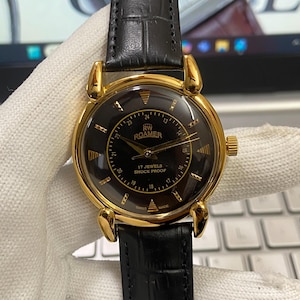 May include: A gold-toned RW Roamer watch with a black face and a black leather band. The watch face features gold-colored hands and markers, with the text "17 Jewels Shock Proof" and "Swiss Made". The watch is held by a gloved hand.