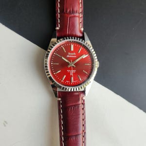 May include: A vintage wristwatch with a red dial and a silver-colored bezel. The watch has a red leather band. The watch face displays the brand name "HMT Kohinoor" and the text "Para Shock 17 Jewels".