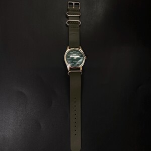 Puede incluir: Un reloj de pulsera con esfera verde y caja plateada. El reloj tiene una correa de cuero verde. La esfera del reloj tiene texto blanco y un peque&ntilde;o logotipo. El reloj se muestra sobre un fondo oscuro.