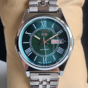 Puede incluir: Un reloj automático Seiko plateado con una esfera verde y marcadores de hora en números romanos. El reloj tiene una ventana de fecha que muestra "SUN 17".