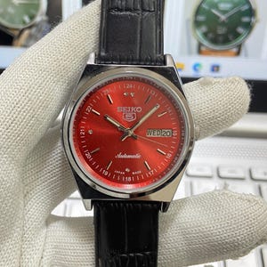 May include: A silver and red Seiko automatic wristwatch with a black leather strap. The watch face has a red background with white markings and a day and date window. The watch face also has the text "SEIKO" and "Automatic" printed on it.