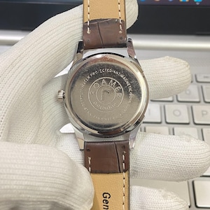 May include: A silver-toned wristwatch with a brown leather band. The watch face is not visible, but the back of the watch has the words "O.A.M.E.R.  WATER PRO. PROTECTED - ANTIMAGNETIC 40 ATM" printed on it.