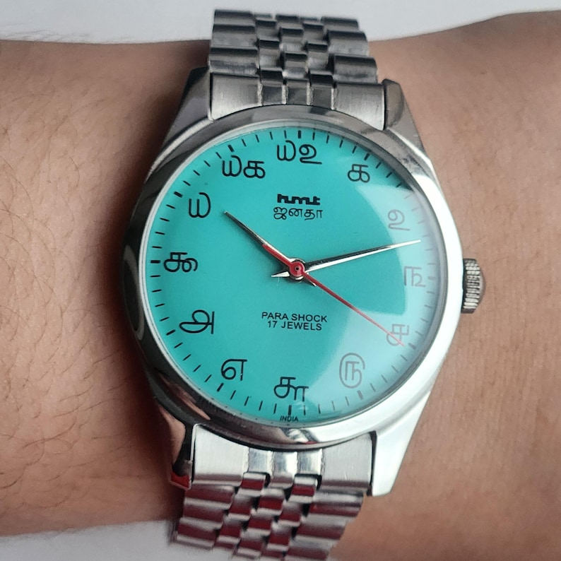 May include: A silver wristwatch with a turquoise face featuring Tamil script numerals and the text "hmt ஜனதா PARA SHOCK 17 JEWELS". The watch has a silver band and red second hand.