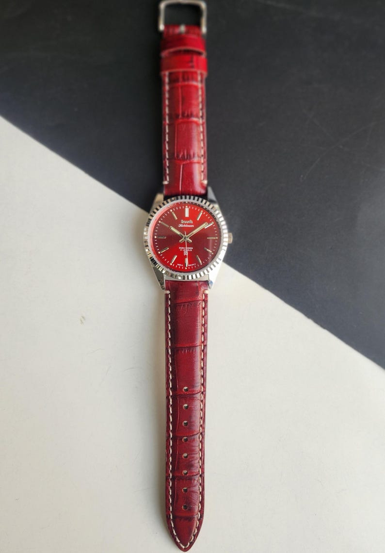 May include: A red wristwatch with a silver bezel and a matching red leather band. The watch face is red with silver hands and markers. The band has a crocodile texture and white stitching.