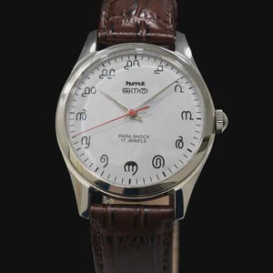 May include: A silver wristwatch with a white face and brown leather strap. The watch face displays unique symbols instead of numbers, with the text "hmt" and "PARA SHOCK 17 JEWELS". The watch has a red second hand.