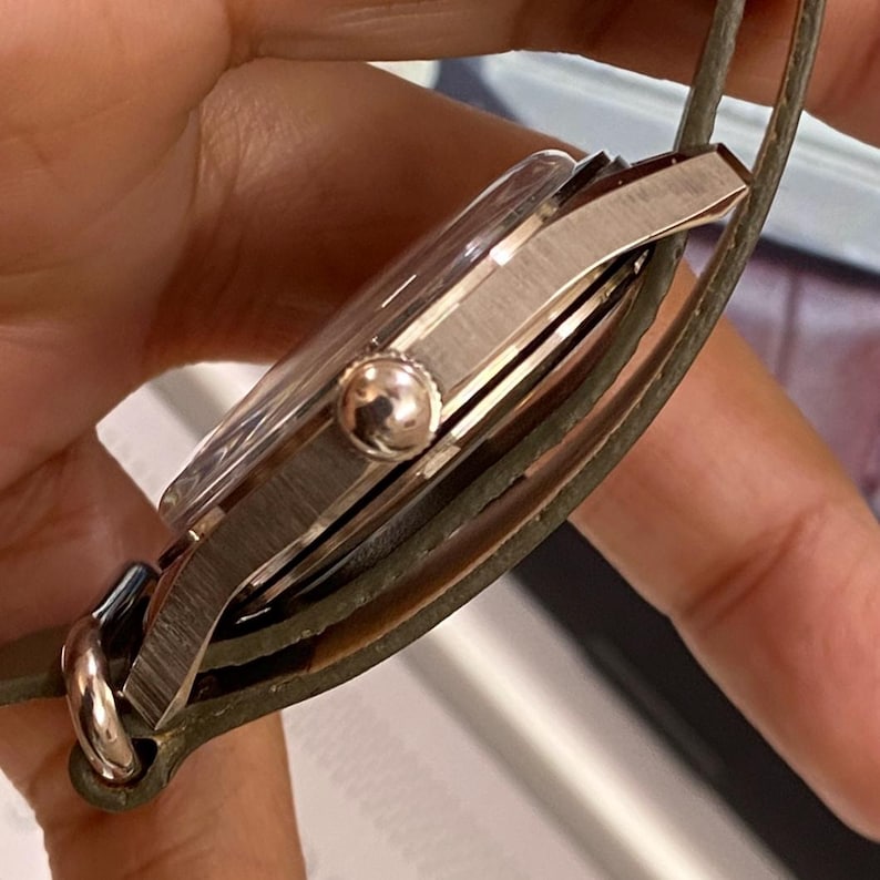 Puede incluir: Un reloj plateado con una esfera de cristal transparente y una corona redonda. El reloj tiene una correa de cuero verde. El reloj est&aacute; sujeto por una mano.